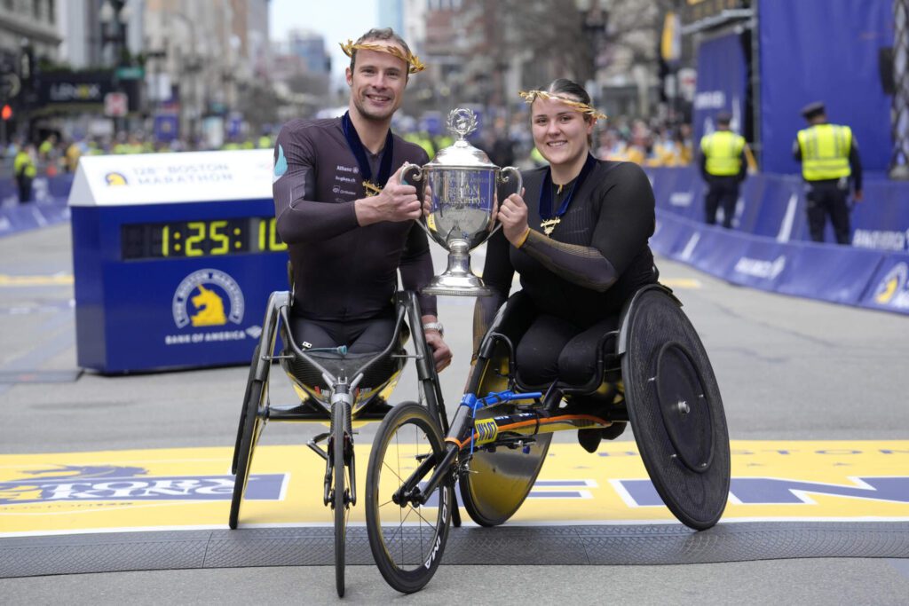 Marcel Hug, da Suíça, à esquerda, vencedor da divisão masculina em cadeiras de rodas da Maratona de Boston, à esquerda, e Eden Rainbow Cooper, da Grã-Bretanha, à direita, vencedor da divisão feminina em cadeiras de rodas, exibem o troféu na linha de chegada em Boston. (Steven Senne/AP)